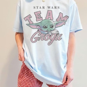Puede incluir: Una camiseta de manga corta de color azul claro con un gr&aacute;fico del personaje Grogu. El texto STAR WARS aparece sobre la palabra TEAM en letras rosas, con la palabra GROGU en letra cursiva rosa debajo de la ilustraci&oacute;n del personaje.