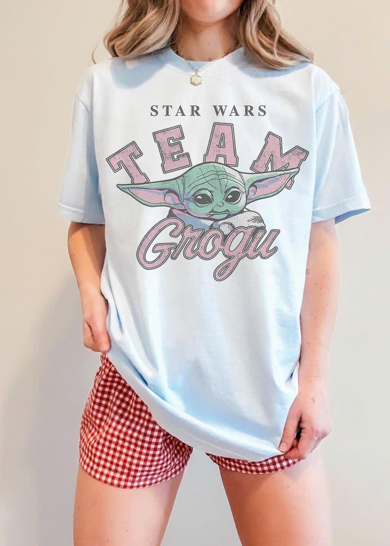 Puede incluir: Una camiseta de manga corta de color azul claro con un gr&aacute;fico del personaje Grogu. El texto STAR WARS aparece sobre la palabra TEAM en letras rosas, con la palabra GROGU en letra cursiva rosa debajo de la ilustraci&oacute;n del personaje.