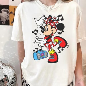 Pu&ograve; includere: T-shirt color crema con un vivace design a cartoni animati di Minnie Mouse. Minnie &egrave; raffigurata mentre balla con note musicali intorno a s&eacute;, indossa un completo rosso con accenti floreali bianchi e tiene in mano un boombox blu. Il design &egrave; giocoso e colorato.