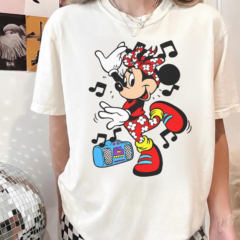 Pu&ograve; includere: T-shirt color crema con un vivace design a cartoni animati di Minnie Mouse. Minnie &egrave; raffigurata mentre balla con note musicali intorno a s&eacute;, indossa un completo rosso con accenti floreali bianchi e tiene in mano un boombox blu. Il design &egrave; giocoso e colorato.