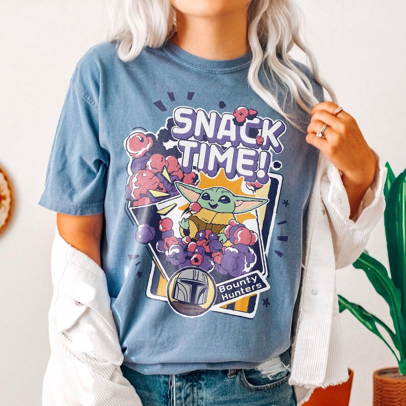 May include: A blue short-sleeved t-shirt featuring a graphic print with the text SNACK TIME! and Bounty Hunters. The design depicts a small green creature with large ears surrounded by purple and pink clouds, with a helmet icon below.