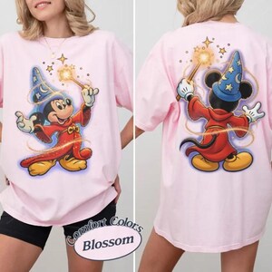 May include: Pale pink t-shirt with a front and back illustration of Mickey Mouse dressed as a sorcerer, holding a wand with stars. The design includes a blue wizard's hat with stars, a red robe, and yellow shoes. The shirt has the text "Comfort Colors Blossom".