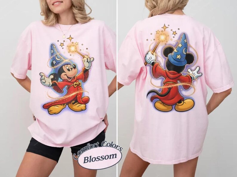 May include: Pale pink t-shirt with a front and back illustration of Mickey Mouse dressed as a sorcerer, holding a wand with stars. The design includes a blue wizard's hat with stars, a red robe, and yellow shoes. The shirt has the text "Comfort Colors Blossom".