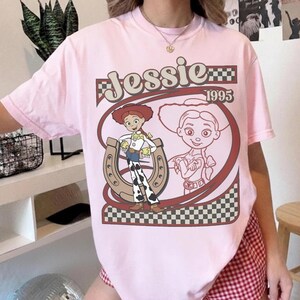 May include: Pink t-shirt featuring a graphic of the character Jessie from Toy Story, with the text "Jessie 1995". The design includes a horseshoe and a checkered pattern, creating a vintage aesthetic. The shirt is a light pink color.