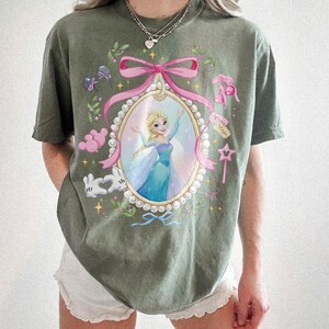 May include: An olive green short-sleeve t-shirt featuring a central oval frame graphic with a pearl border and a pink ribbon. Inside the frame is a blonde character in a blue dress. The design includes white gloves, pink balloons, and a star wand.