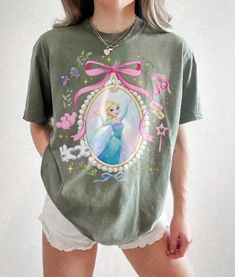 May include: An olive green short-sleeve t-shirt featuring a central oval frame graphic with a pearl border and a pink ribbon. Inside the frame is a blonde character in a blue dress. The design includes white gloves, pink balloons, and a star wand.