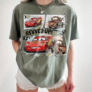 May include: An olive green t-shirt featuring a graphic print of the characters Lightning McQueen and Mater from the movie Cars. The design includes comic-style panels, the text REVVED UP!, and checkered flag accents in red, brown, and black.