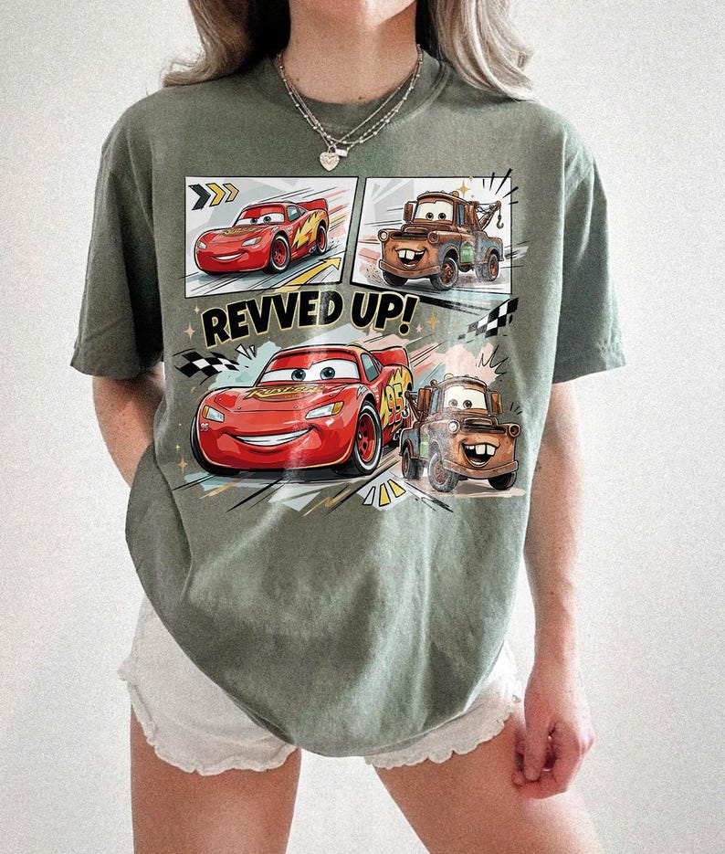 May include: An olive green t-shirt featuring a graphic print of the characters Lightning McQueen and Mater from the movie Cars. The design includes comic-style panels, the text REVVED UP!, and checkered flag accents in red, brown, and black.