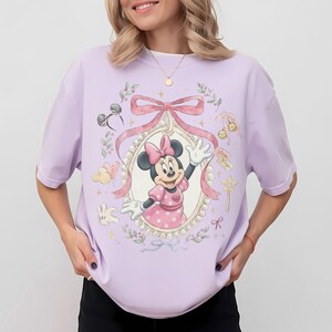 May include: A light purple t-shirt featuring a central graphic of Minnie Mouse inside an ornate, pearl-bordered oval frame. The design includes a large pink bow, floral accents, small star sparkles, a wand, and mouse ear headbands in soft pastel tones.