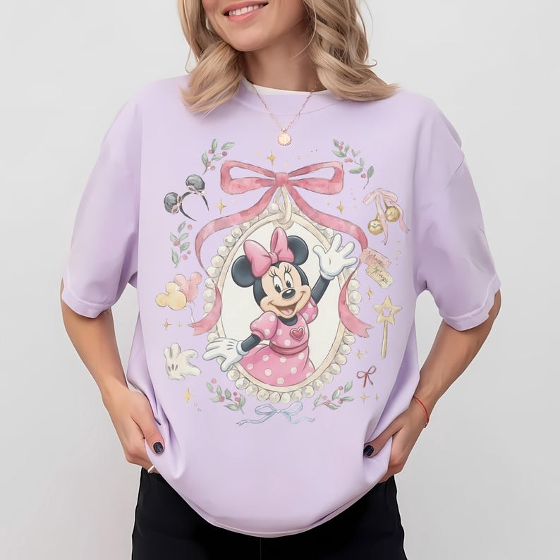 May include: A light purple t-shirt featuring a central graphic of Minnie Mouse inside an ornate, pearl-bordered oval frame. The design includes a large pink bow, floral accents, small star sparkles, a wand, and mouse ear headbands in soft pastel tones.