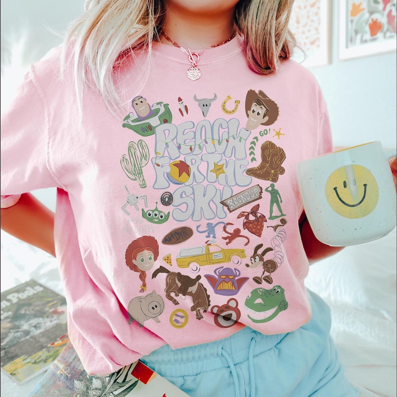 May include: A light pink t-shirt featuring a graphic print with the text "REACH FOR THE SKY" surrounded by various cartoon character illustrations, including a cowboy, a space ranger, a cowgirl, a dinosaur, a pig, and a yellow truck.