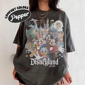 Puede incluir: Camiseta gris oscuro con un gr&aacute;fico de Disneylandia con Mickey Mouse, Minnie Mouse, Donald Duck y otros personajes de Disney. La camiseta tiene el texto "Disneyland Est. 1955" y "Comfort Colors Pepper".