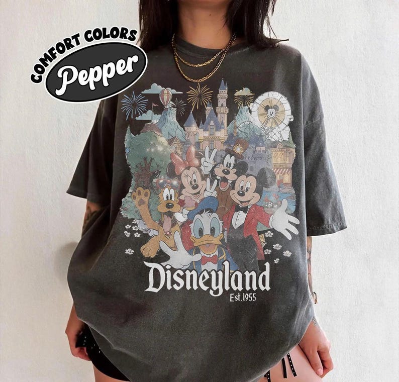 Puede incluir: Camiseta gris oscuro con un gr&aacute;fico de Disneylandia con Mickey Mouse, Minnie Mouse, Donald Duck y otros personajes de Disney. La camiseta tiene el texto "Disneyland Est. 1955" y "Comfort Colors Pepper".