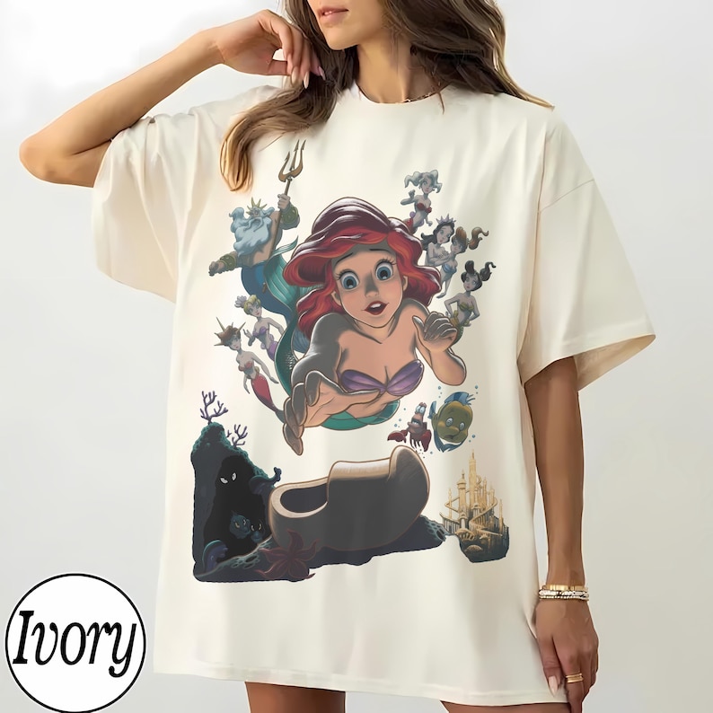 May include: An ivory t-shirt featuring a large, colorful graphic print of a red-haired mermaid character surrounded by various underwater figures, including a crab, a fish, a king with a trident, and a castle, with the word Ivory in a circle.