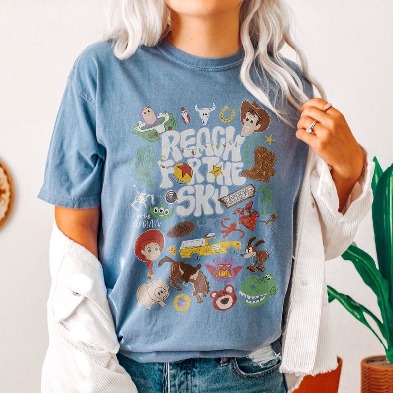 May include: A blue short-sleeve t-shirt featuring a graphic print with the text "REACH FOR THE SKY" surrounded by various cartoon character illustrations, including a cowboy, a space ranger, a dinosaur, a pig, and a yellow truck in a collage style.