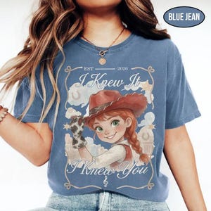 May include: A blue jean coloured t-shirt featuring a graphic of a cartoon cowgirl wearing a brown hat and a cow-print glove. The text reads EST 2026, I Knew It, and I Knew You, framed by a rope design with stars and horseshoes in the background.
