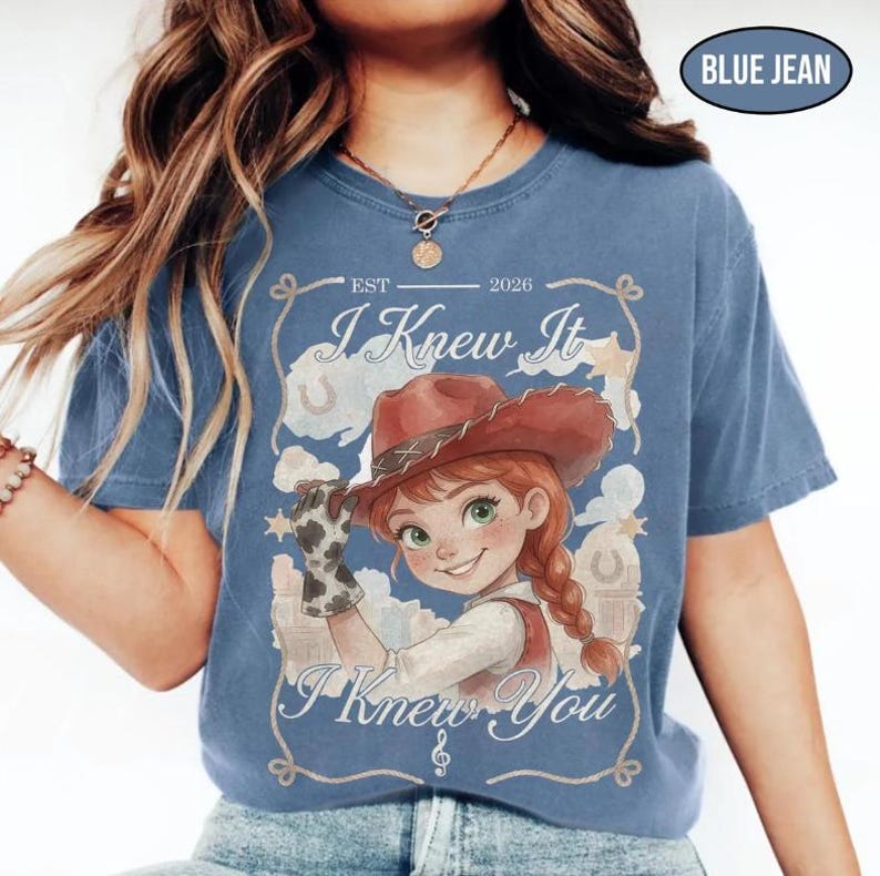May include: A blue jean coloured t-shirt featuring a graphic of a cartoon cowgirl wearing a brown hat and a cow-print glove. The text reads EST 2026, I Knew It, and I Knew You, framed by a rope design with stars and horseshoes in the background.