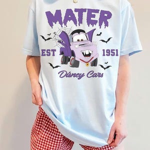 May include: Light blue t-shirt featuring a cartoon Mater character in a vampire costume, with the text "MATER EST 1951 Disney Cars". The design includes bats and a spiderweb. The shirt is worn with red and white checkered shorts.