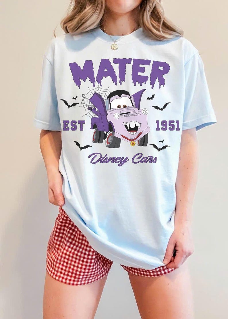 May include: Light blue t-shirt featuring a cartoon Mater character in a vampire costume, with the text "MATER EST 1951 Disney Cars". The design includes bats and a spiderweb. The shirt is worn with red and white checkered shorts.