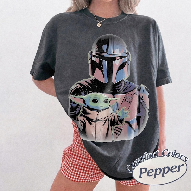 May include: Dark gray t-shirt featuring a graphic of The Mandalorian and Grogu. The Mandalorian is in a helmet and armor, holding Grogu. The shirt has the words "Comfort Colors Pepper" in a circular design.