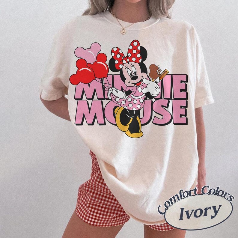 May include: Ivory-colored t-shirt featuring a graphic of Minnie Mouse holding balloons and an ice cream cone. The text "MINNIE MOUSE" is in pink block letters. The shirt is labeled "Comfort Colors Ivory".