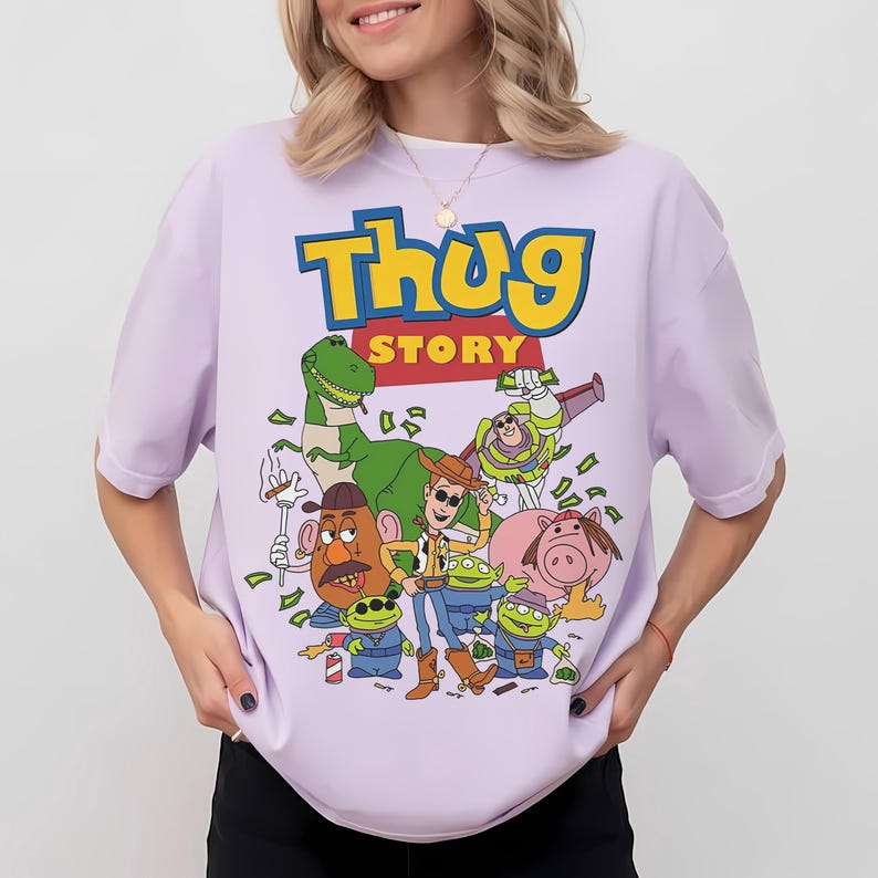 Pode incluir: Camiseta lil&aacute;s claro com um gr&aacute;fico de desenho animado com o texto "Thug Story" em amarelo e azul. O gr&aacute;fico inclui v&aacute;rios personagens de desenho animado e notas de dinheiro.