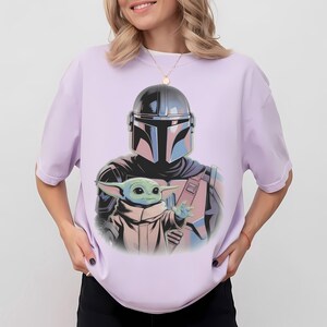 May include: Lilac-colored t-shirt featuring a graphic of the Mandalorian and Grogu. The Mandalorian is depicted in his helmet and armor, holding Grogu. The artwork has a comic book style with a color palette of black, blue, pink, and green.