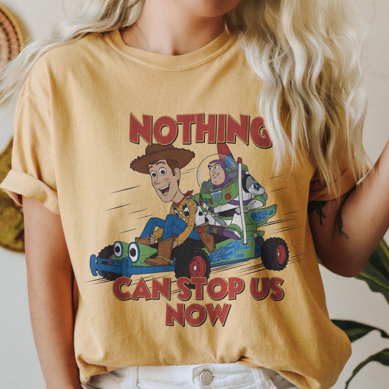 以下が含まれることがあります： マスタードイエローのTシャツ。トイ・ストーリーのウッディとバズ・ライトイヤーがレーシングカーに乗ったグラフィック。テキスト「NOTHING CAN STOP US NOW」は赤色で印刷されています。Tシャツは半袖で、クルーネックです。