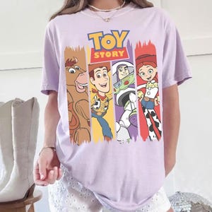 May include: A light purple t-shirt featuring the words "TOY STORY" in blue and yellow. The shirt displays four cartoon characters in vertical color blocks: a brown horse, Woody, Buzz Lightyear, and Jessie. The shirt is short-sleeved.