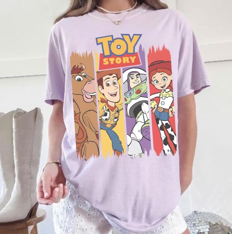 May include: A light purple t-shirt featuring the words "TOY STORY" in blue and yellow. The shirt displays four cartoon characters in vertical color blocks: a brown horse, Woody, Buzz Lightyear, and Jessie. The shirt is short-sleeved.