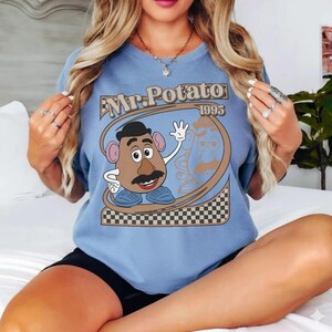May include: A light blue t-shirt featuring a graphic of Mr. Potato Head with the text "Mr. Potato 1995". The design includes a cartoon image of Mr. Potato Head with a brown mustache, black hat, and blue shoes. The shirt has a checkered pattern at the bottom.