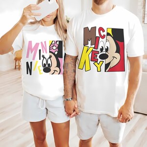 Op de afbeelding: Twee witte T-shirts met cartoonafbeeldingen. E&eacute;n shirt heeft "MINNIE" in roze, zwart en geel met Minnie Mouse. Het andere shirt toont "MICKEY" in bruin en geel met Mickey Mouse. Beide shirts worden gedragen met lichtgrijze shorts.