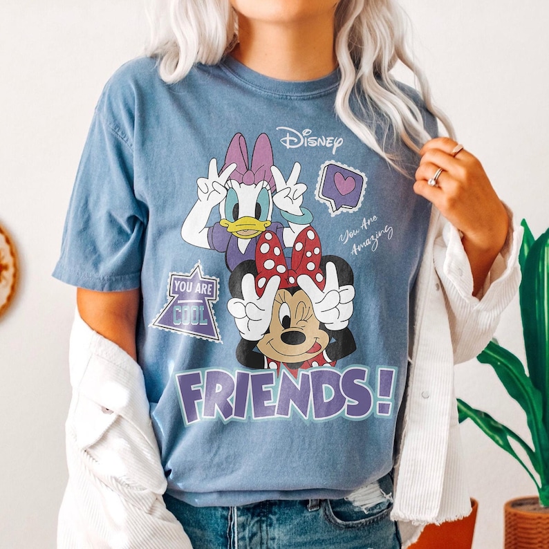 Może przedstawiać: Niebieska koszulka z Myszką Minnie i Daisy Duck pokazującymi znak pokoju. Koszulka ma napis "FRIENDS!" w fioletowych literach oraz frazy "You Are Cool" i "You Are Amazing". Logo Disneya znajduje się na g&oacute;rze.