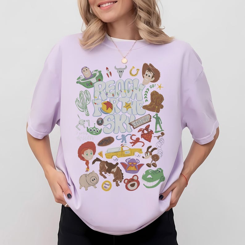 May include: A light purple t-shirt featuring a graphic print with the text "REACH FOR THE SKY" surrounded by various cartoon character illustrations, including a space ranger, a cowboy, a cowgirl, a green alien, a pig, a dinosaur, and a yellow truck.