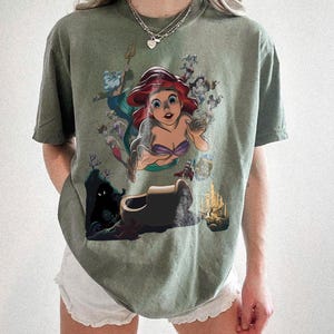 May include: An olive green short-sleeve t-shirt featuring a graphic print of the character Ariel from The Little Mermaid, surrounded by other characters including King Triton, Sebastian, and Flounder, along with an underwater castle and cave.