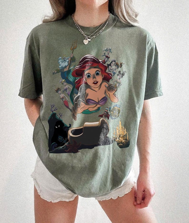 May include: An olive green short-sleeve t-shirt featuring a graphic print of the character Ariel from The Little Mermaid, surrounded by other characters including King Triton, Sebastian, and Flounder, along with an underwater castle and cave.