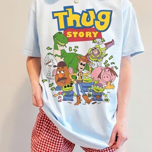 Pode incluir: Camiseta azul clara com um gr&aacute;fico de desenho animado e o texto "Thug Story" em amarelo e azul. O gr&aacute;fico inclui v&aacute;rios personagens de desenho animado. A pessoa est&aacute; usando shorts xadrez vermelhos e brancos.