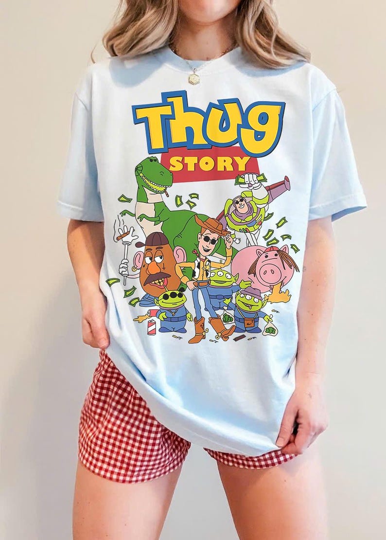 Pode incluir: Camiseta azul clara com um gr&aacute;fico de desenho animado e o texto "Thug Story" em amarelo e azul. O gr&aacute;fico inclui v&aacute;rios personagens de desenho animado. A pessoa est&aacute; usando shorts xadrez vermelhos e brancos.