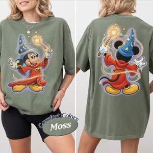 May include: An olive green t-shirt with a cartoon Mickey Mouse wizard design on the front and back. The character is holding a wand with a glowing star. The shirt has the words "Comfort Colors Moss" on the bottom.