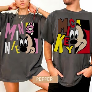 Op de afbeelding: Twee antracietgrijze t-shirts met stripfiguren. E&eacute;n shirt toont "MINNIE" in roze, wit en geel, met het gezicht van Minnie Mouse. Het andere shirt toont "MICKEY" in bruin en geel, met het gezicht van Mickey Mouse. Beide shirts hebben een relaxte pasvorm.