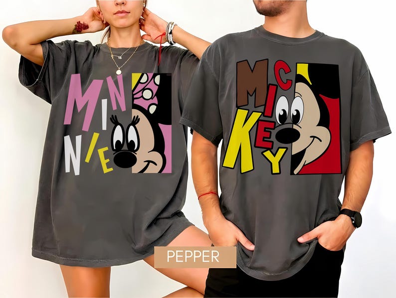 Op de afbeelding: Twee antracietgrijze t-shirts met stripfiguren. E&eacute;n shirt toont "MINNIE" in roze, wit en geel, met het gezicht van Minnie Mouse. Het andere shirt toont "MICKEY" in bruin en geel, met het gezicht van Mickey Mouse. Beide shirts hebben een relaxte pasvorm.