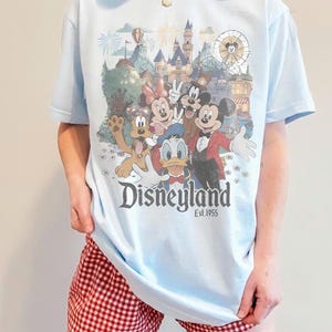 Puede incluir: Camiseta azul claro con un dise&ntilde;o de Disneyland con Mickey Mouse, Minnie Mouse, Donald Duck y Goofy. La camiseta tiene el texto "Disneyland Est.1955". La persona lleva un pantal&oacute;n corto a cuadros rojos y blancos.