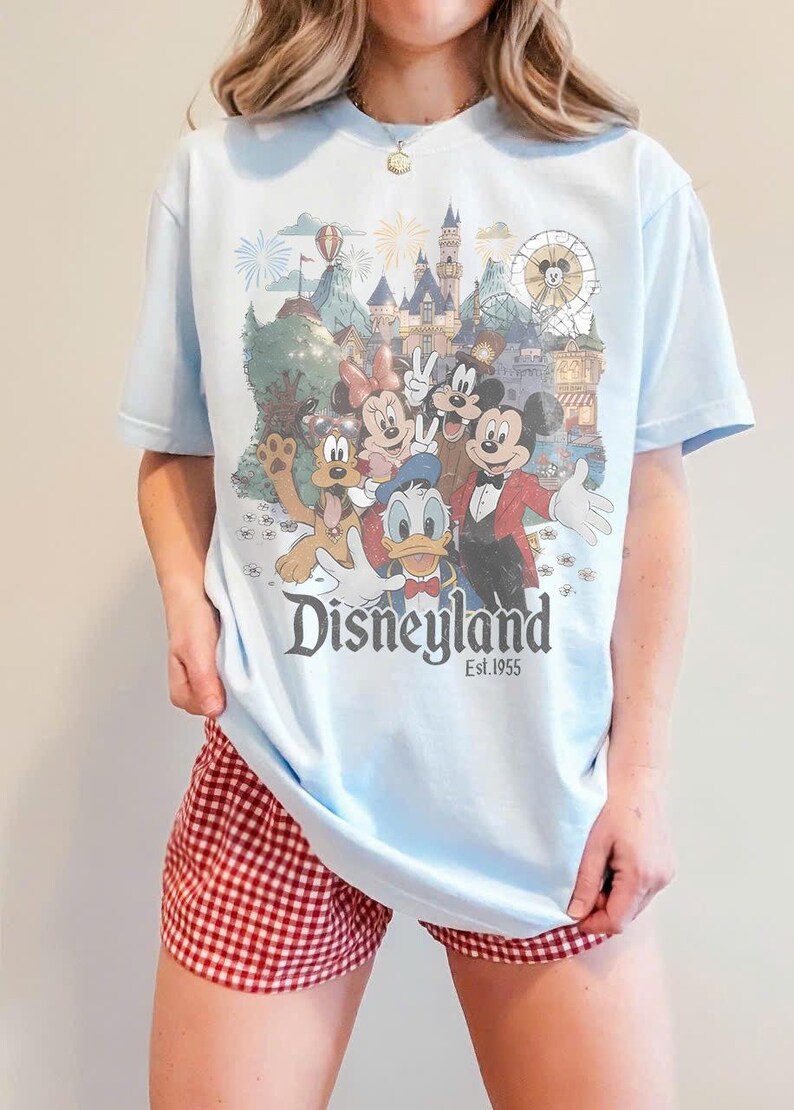 Puede incluir: Camiseta azul claro con un dise&ntilde;o de Disneyland con Mickey Mouse, Minnie Mouse, Donald Duck y Goofy. La camiseta tiene el texto "Disneyland Est.1955". La persona lleva un pantal&oacute;n corto a cuadros rojos y blancos.