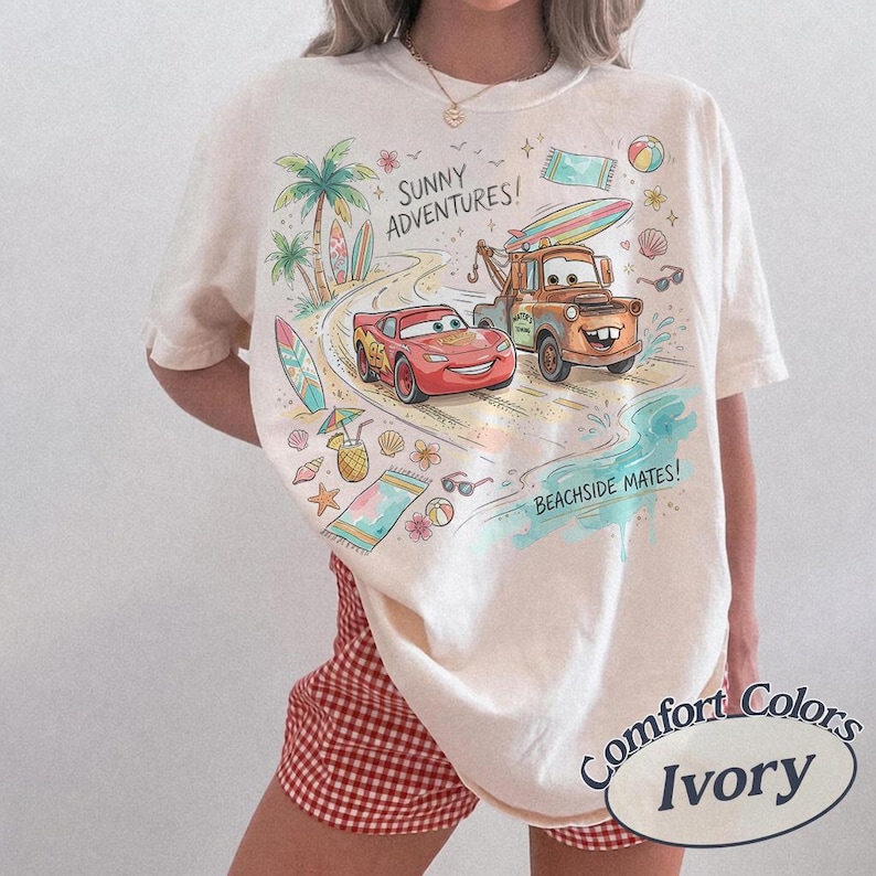 May include: An ivory t-shirt featuring a colorful graphic of Lightning McQueen and Mater driving on a beach road. The design includes palm trees, surfboards, beach towels, and the text "SUNNY ADVENTURES!" and "BEACHSIDE MATES!" in a playful font.