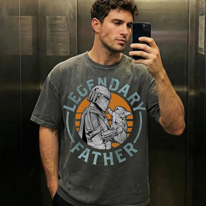 K&ouml;nnte beinhalten: Graues T-Shirt mit einer Grafik von The Mandalorian und Grogu, mit dem Text "LEGENDARY FATHER" in Blau und Wei&szlig;. Das Design zeigt einen orangefarbenen Kreis hinter den Figuren. Das Shirt ist ein l&auml;ssiger Stil.