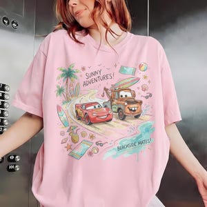 May include: A light pink t-shirt featuring a colorful graphic of Lightning McQueen and Mater from the movie Cars on a beach road. The design includes palm trees, surfboards, beach towels, and the text "SUNNY ADVENTURES!" and "BEACHSIDE MATES!" printed in a playful font.