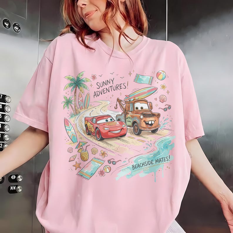 May include: A light pink t-shirt featuring a colorful graphic of Lightning McQueen and Mater from the movie Cars on a beach road. The design includes palm trees, surfboards, beach towels, and the text "SUNNY ADVENTURES!" and "BEACHSIDE MATES!" printed in a playful font.