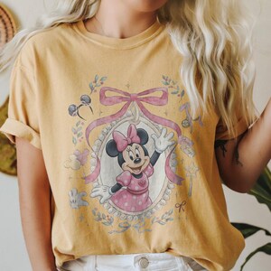May include: A mustard yellow short-sleeve t-shirt featuring a graphic print of Minnie Mouse inside an oval frame decorated with a large pink bow. The design includes small illustrations of mouse ears, balloons, a star wand, and floral accents.