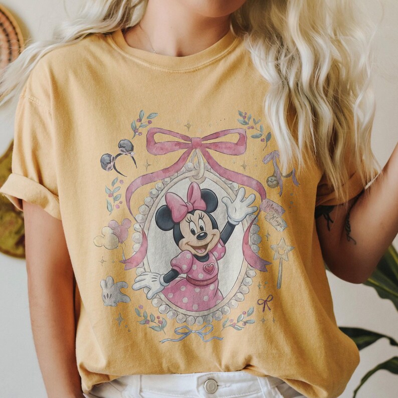 May include: A mustard yellow short-sleeve t-shirt featuring a graphic print of Minnie Mouse inside an oval frame decorated with a large pink bow. The design includes small illustrations of mouse ears, balloons, a star wand, and floral accents.