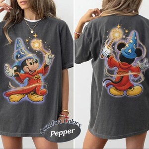 May include: Dark grey t-shirt with a front and back print of Mickey Mouse as a sorcerer, wearing a blue hat and red robe, holding a wand. The shirt has a distressed appearance and the text "Comfort Colors Pepper" is printed on the bottom.